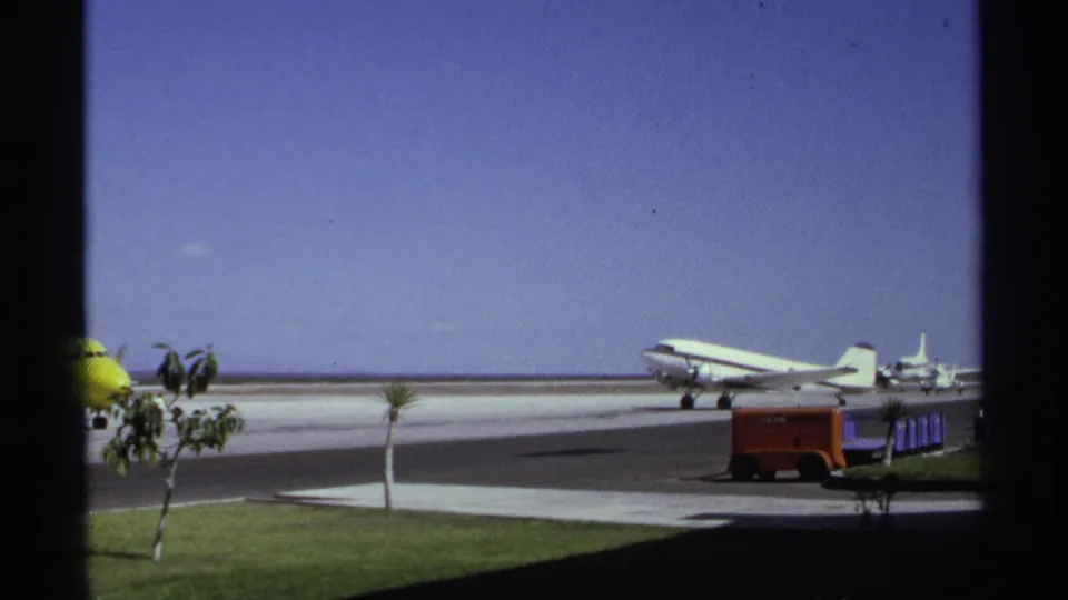 1974 La Paz airport frame showing a white propeller aircraft, red ground equipment, palms, grass, and the yellow plane partly at left.