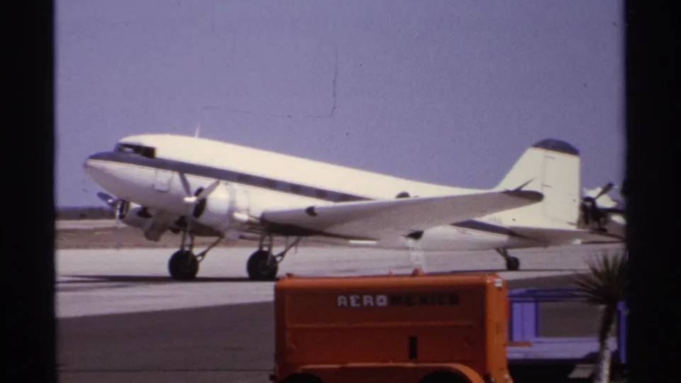 Close 8mm frame from La Paz, Mexico in 1974 showing a white propeller airliner and orange Aero Mexico vehicle on the airport apron.