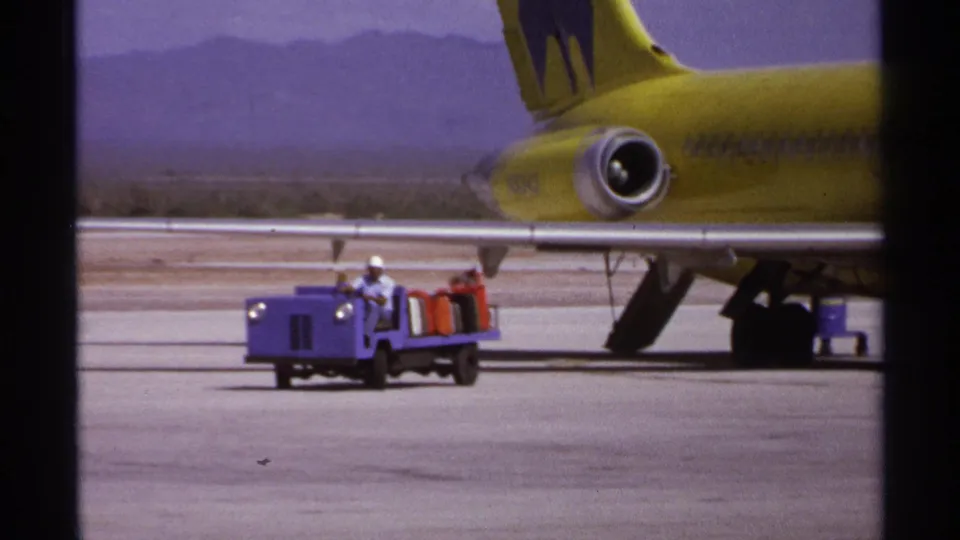 1974 La Paz airport scene with a yellow jet parked near distant mountains while a small purple luggage cart carries bags and ground crew.