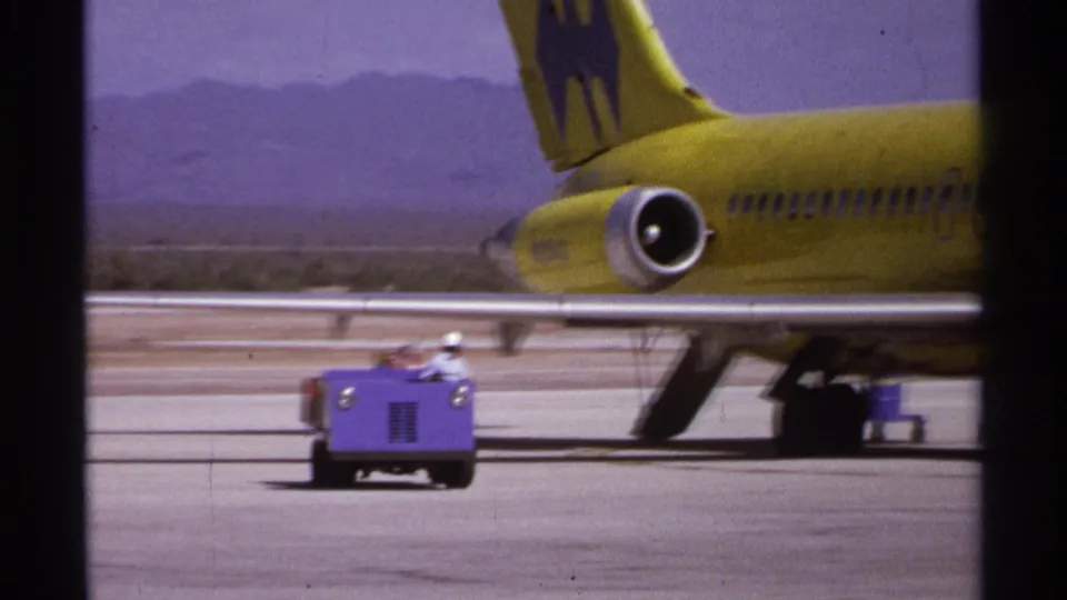 A small purple baggage cart moves across the La Paz tarmac in front of a yellow jet, with mountains and runway space in the background.