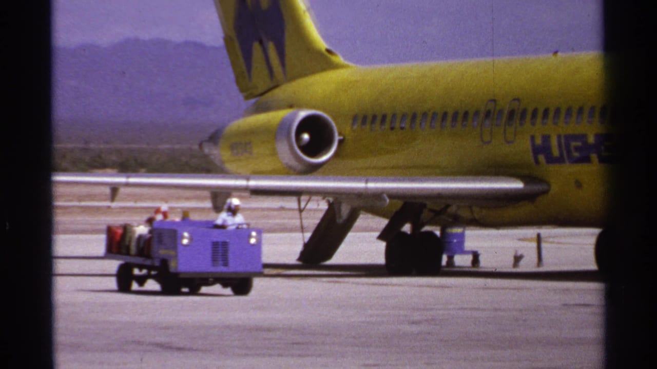Yellow Hughes Airwest plane tail and rear fuselage beside a purple baggage tug on the tarmac.
