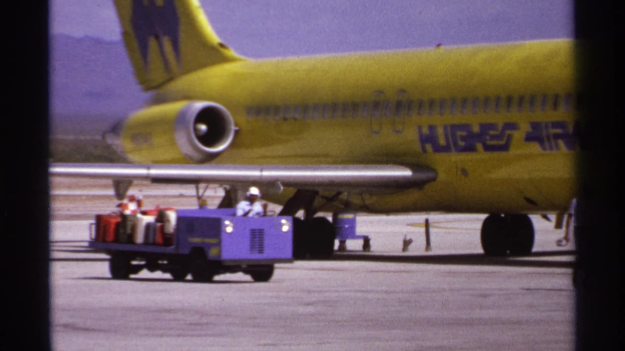 Purple baggage tug with suitcases passes in front of a yellow Hughes Airwest aircraft.