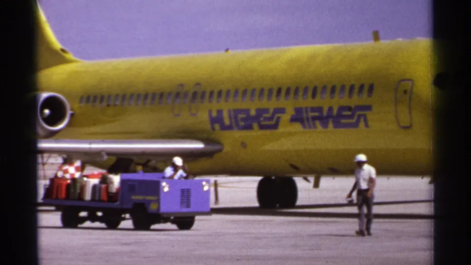 1974 La Paz tarmac view with the Hughes Airwest name on the fuselage, baggage equipment below, and a hard-hatted worker walking through frame.