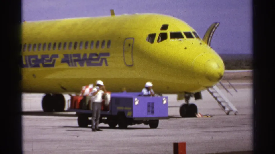 1974 airport ground crew activity in La Paz, with a yellow Hughes Airwest aircraft, baggage cart, suitcases, and boarding stairs visible.