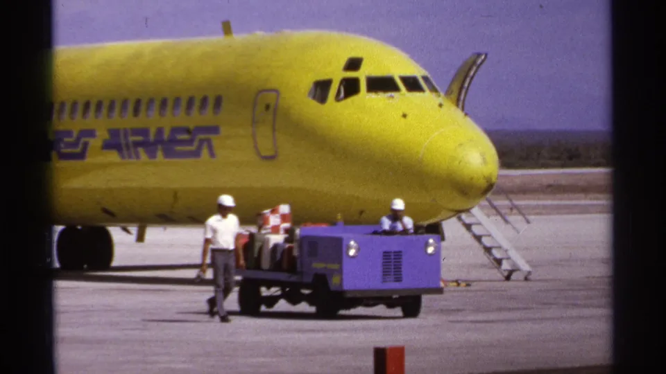 La Paz 1974 home-movie frame showing a baggage cart and workers beneath the nose of a yellow Hughes Airwest plane.