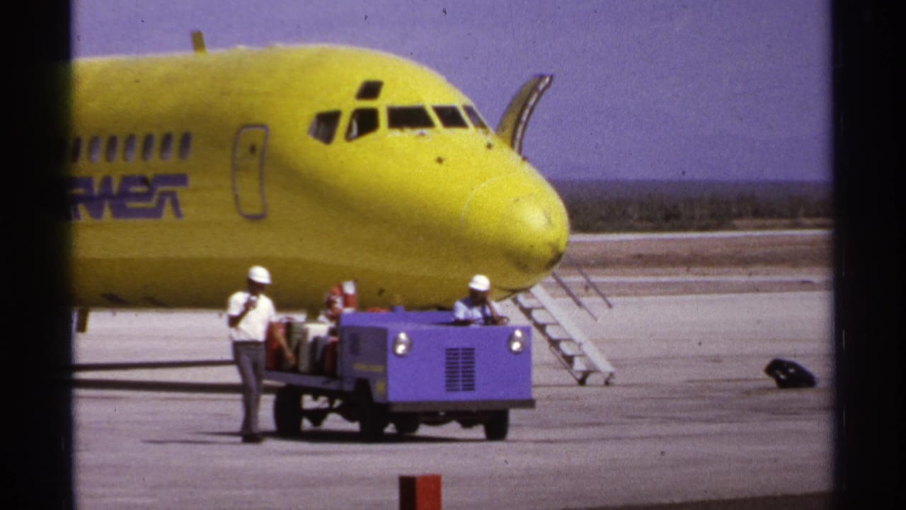 1974 La Paz, Mexico tarmac view with a yellow airliner, hard-hatted ground crew, and luggage being moved on a purple cart.