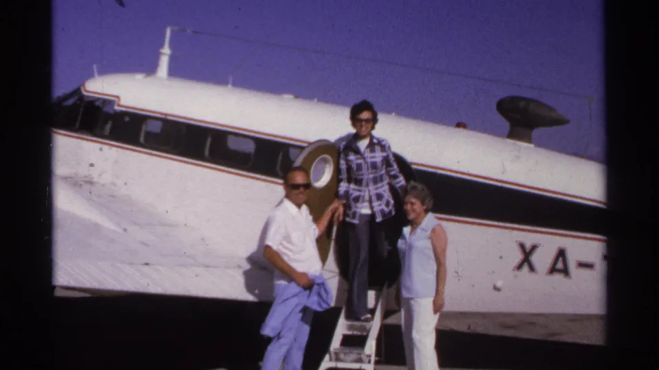 Three adults gather at the steps of a small white propeller aircraft in 1974 La Paz, Mexico home-movie footage.
