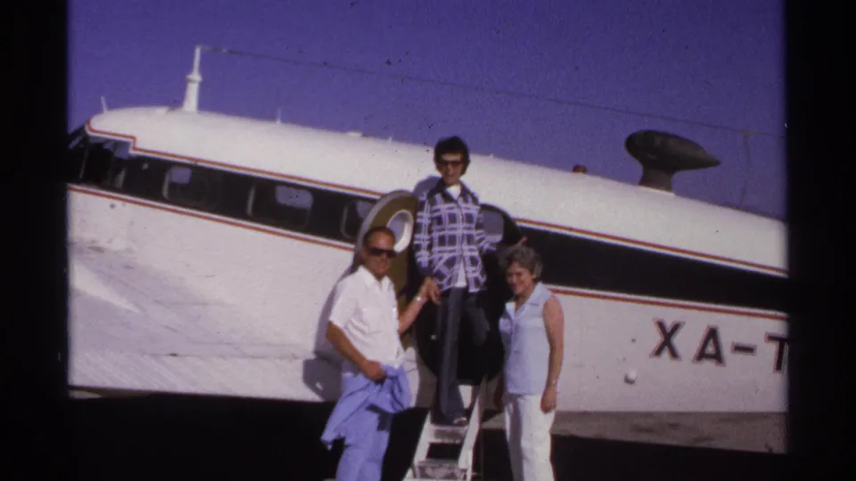 Adults wave and pose beside a small white propeller aircraft marked with partial XA registration in La Paz, Mexico, 1974.