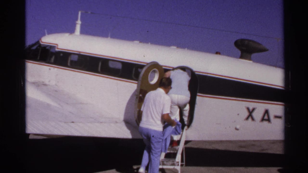 Two men stand at portable stairs beside a small white aircraft on a sunny tarmac.
