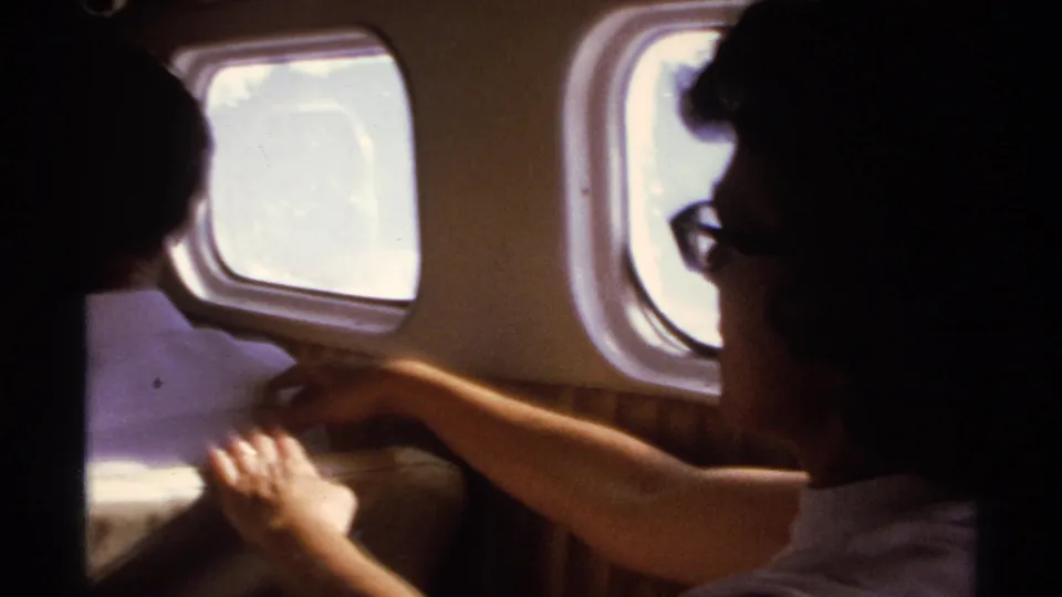 Interior 1974 La Paz travel frame showing seated adult passengers in a small aircraft cabin with bright windows behind them.