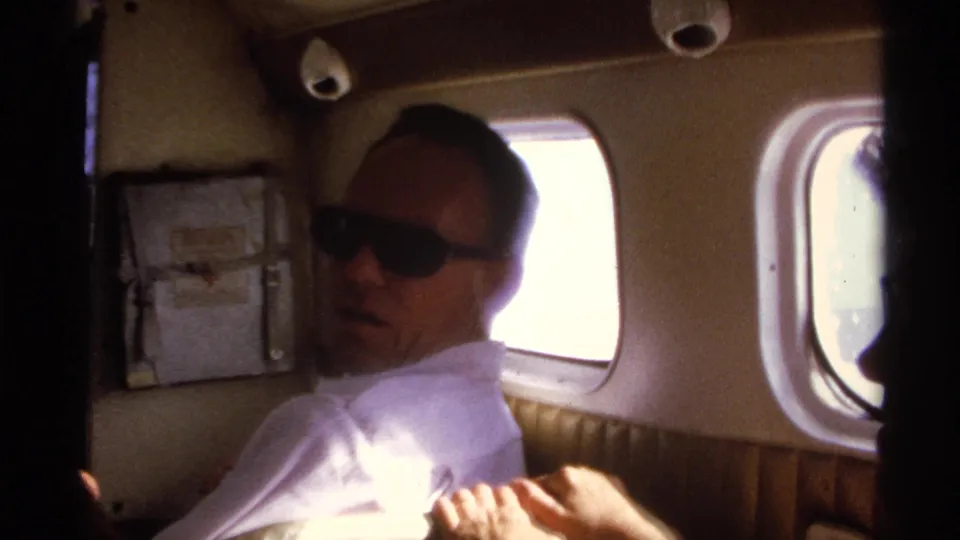 1974 La Paz home-movie frame of an adult man in sunglasses seated in a small aircraft cabin beside bright windows.