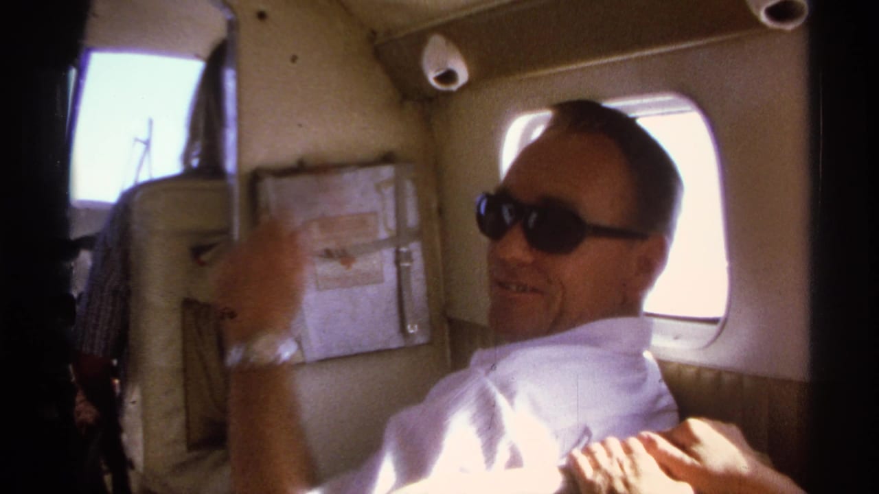 Man in sunglasses waves from his seat beside rounded aircraft windows.