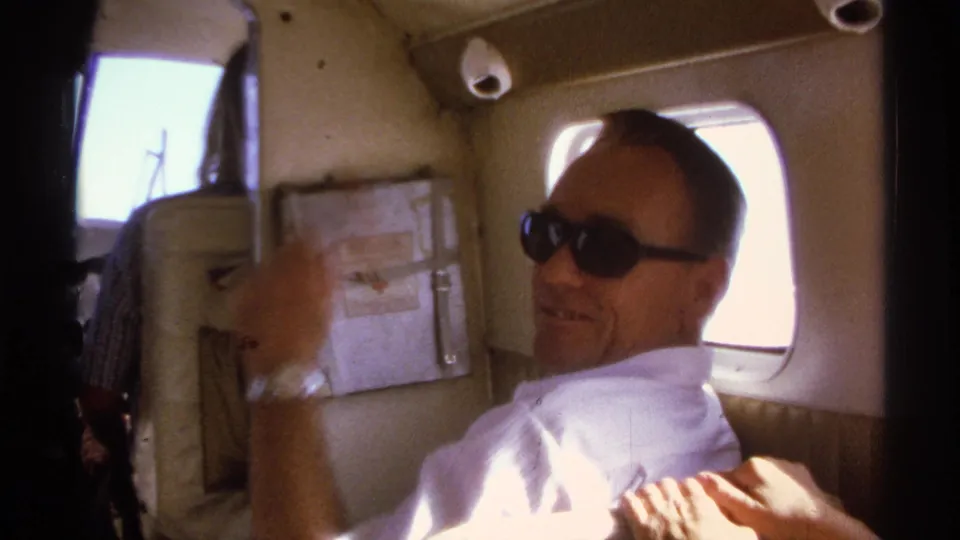 The passenger raises a hand in a wave while seated beside rounded windows in the small aircraft cabin.