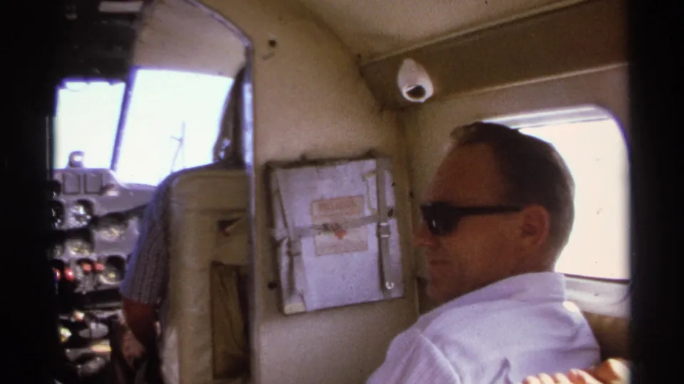 The man turns away from the camera, with a cockpit area, seat backs, and analog controls visible ahead in the aircraft.