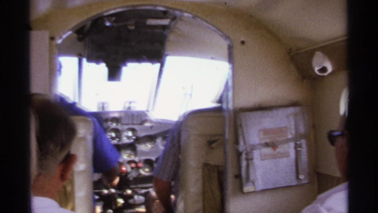 Cockpit view from behind the seats with men near the controls and bright windshield.