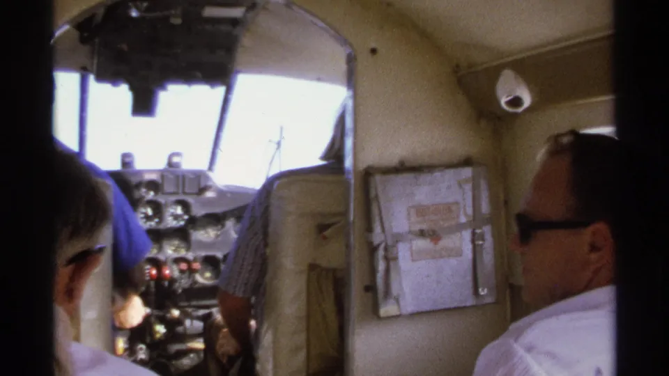 Handheld 8mm frame from inside a small aircraft cabin, with men near the instrument panel and sunlight through the windshield.