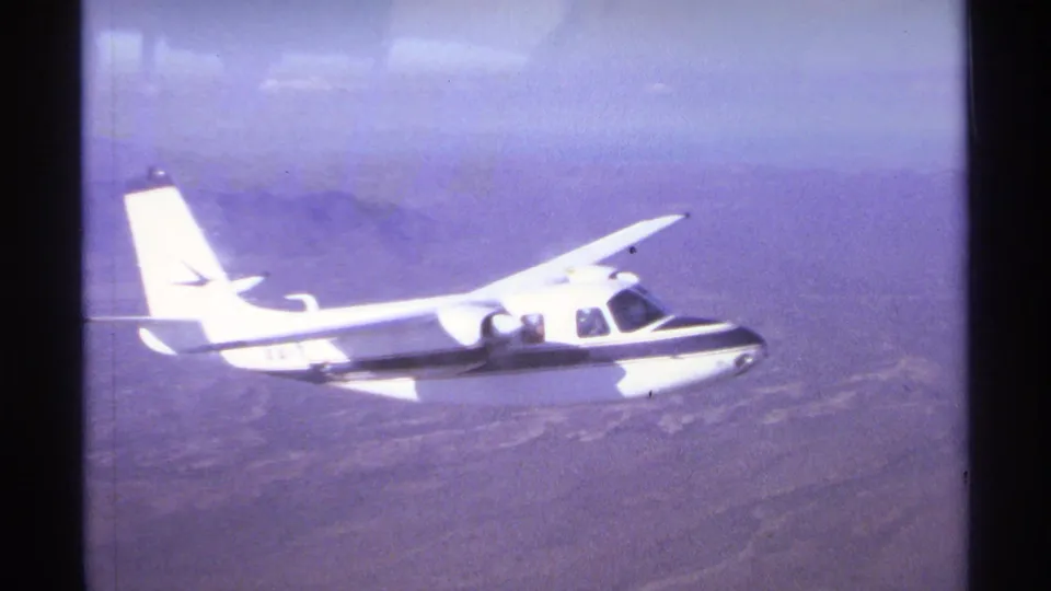 1974 home-movie frame showing a white small aircraft in flight over hazy dry landscape near La Paz, Mexico.