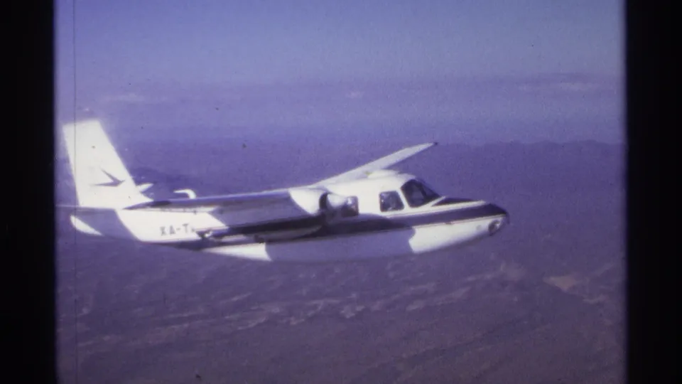 The small white aircraft remains in profile over an arid landscape, with 8mm grain and faded color visible.