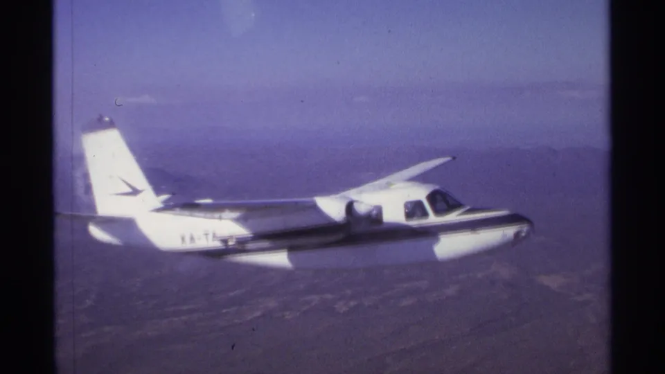 A 1974 8mm frame shows a white aircraft with partial XA-T marking flying over dry terrain near La Paz, Mexico.
