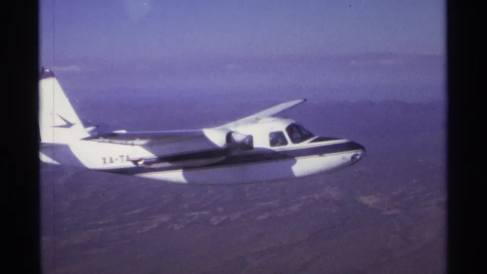 1974 La Paz home-movie frame showing a small white propeller plane with dark stripe above a hazy, dry landscape.