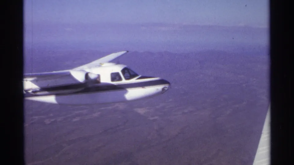 White-and-dark propeller plane filmed from another aircraft, with arid land and distant mountains visible below.