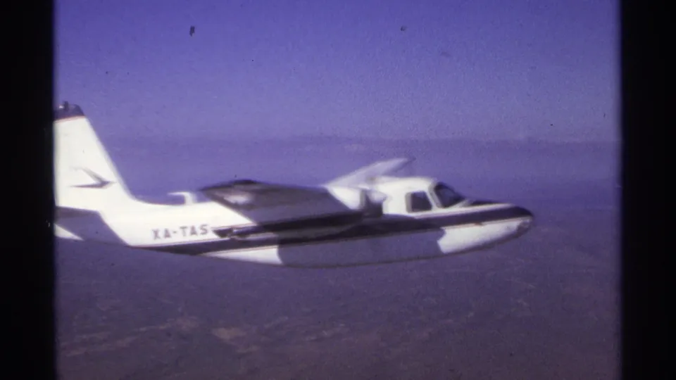 1974 8mm frame near La Paz, Mexico, showing the side of a small propeller plane with partial tail markings over arid terrain.