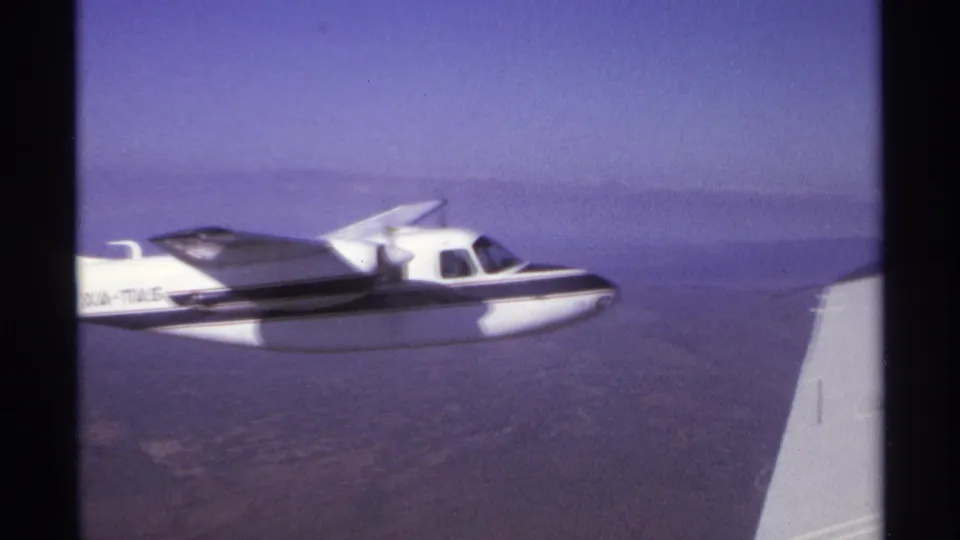 1974 8mm frame near La Paz, Mexico, showing a small propeller plane passing the filming aircraft wing over dry terrain.