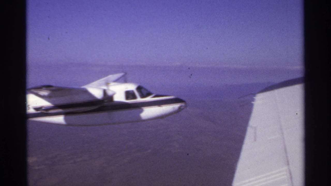 Small white aircraft flying beside a visible aircraft wing over hazy water.