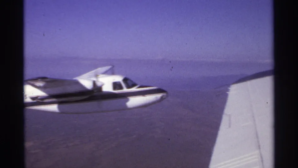 1974 La Paz, Mexico 8mm frame showing a small white aircraft with a dark stripe flying near the filming aircraft wing.
