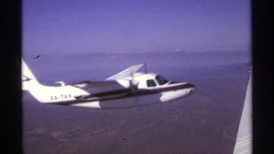 The XA-TAS aircraft is framed lower beside the filming aircraft wing, with hazy water and sky in the background.