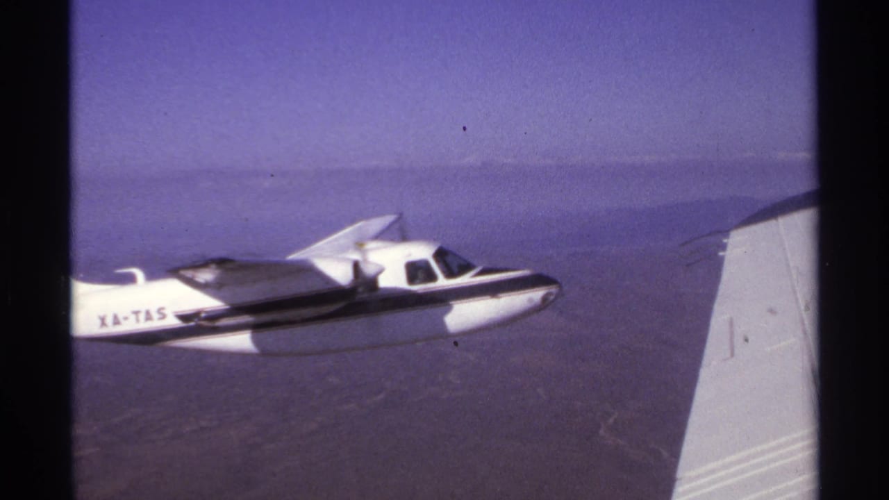 1974 8mm frame near La Paz, Mexico showing a white single-engine airplane marked XA-TAS flying over rugged terrain.