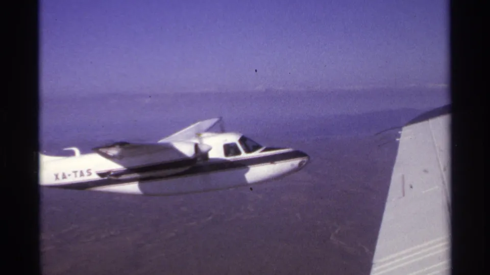 1974 8mm frame near La Paz, Mexico showing a white single-engine airplane marked XA-TAS flying over rugged terrain.