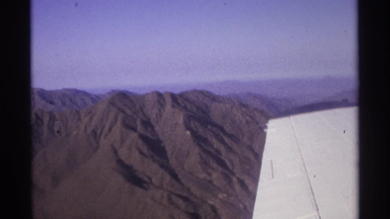 Aircraft wing in the foreground with rugged mountains below and a hazy horizon.
