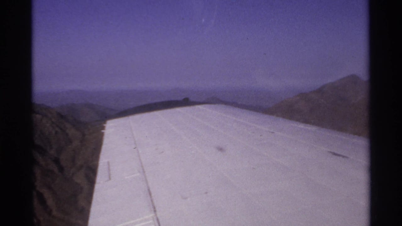 White airplane wing seen over rugged mountains through a dark-framed aircraft window.