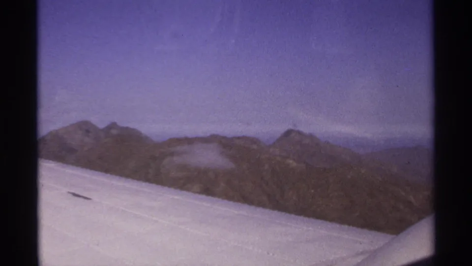 1974 home-movie frame from a plane over La Paz, Mexico, showing a wing, mountain range, and hazy sky with visible film wear.