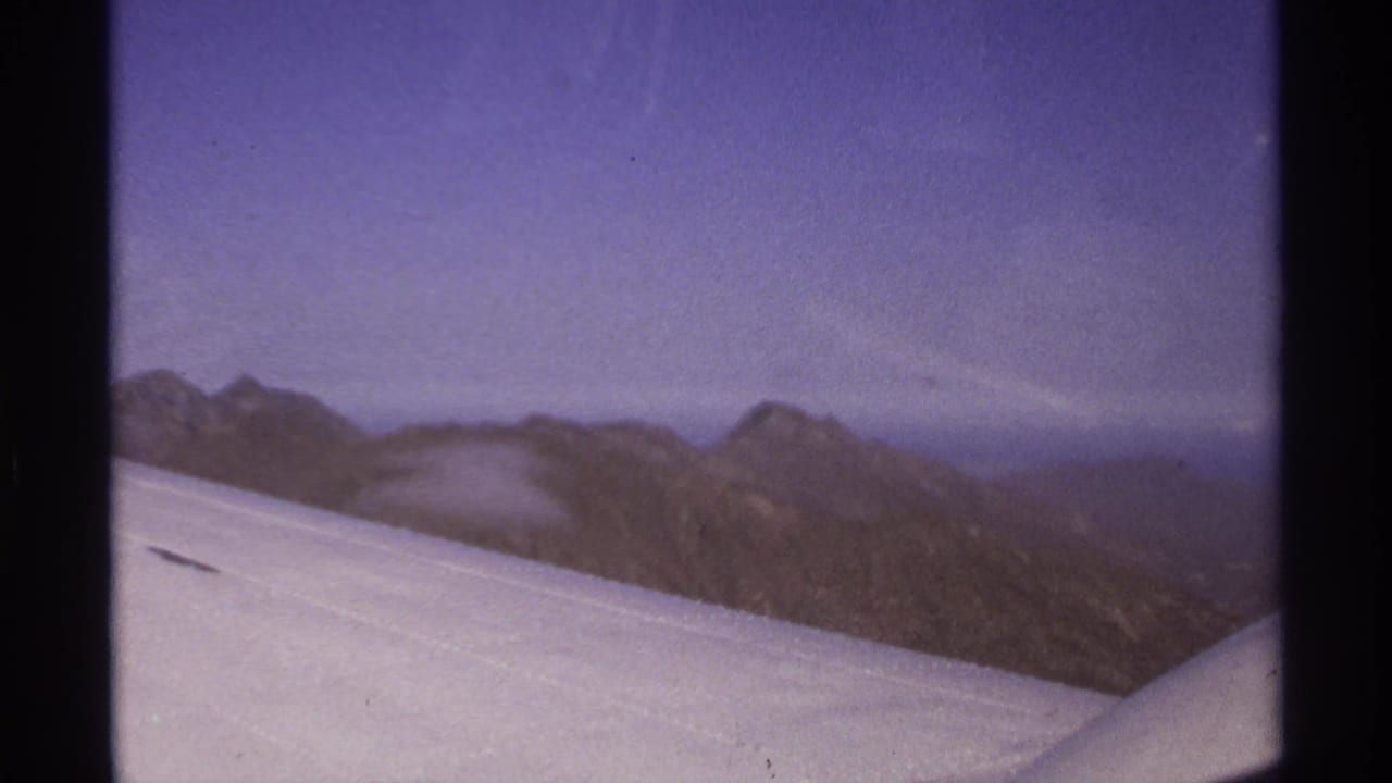 Aircraft wing and window frame over rugged mountains under a hazy sky.