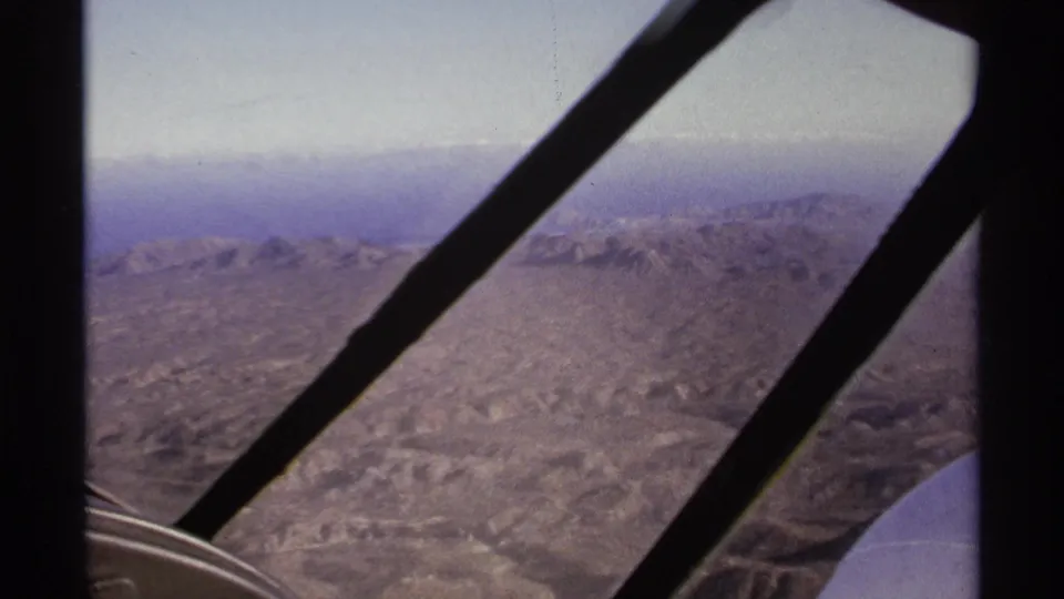 1974 home-movie frame of arid mountains near La Paz, Mexico, viewed through dark airplane window bars and faded small-gauge color.