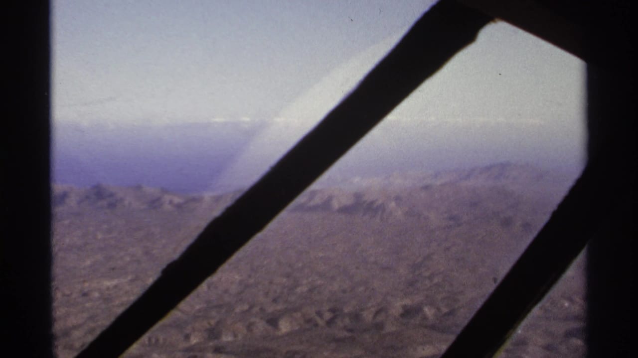 1974 8mm aerial view near La Paz, Mexico, with dark aircraft window supports crossing hazy mountain ridges and desert valleys.