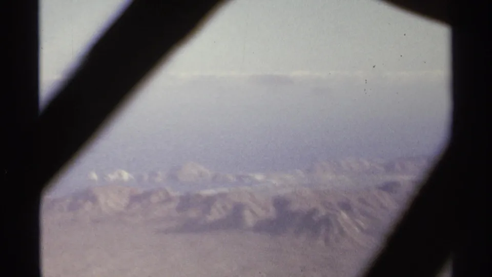 Soft 1974 8mm aerial view near La Paz, with dark window framing, hazy terrain, and pale distant summits beyond the desert ridges.