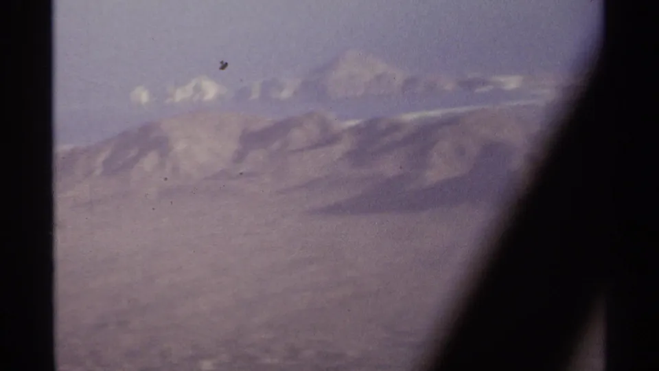Final aerial frame from the 1974 La Paz, Mexico clip, showing desert mountain ridges, pale distant summits, and a dark aircraft window edge.