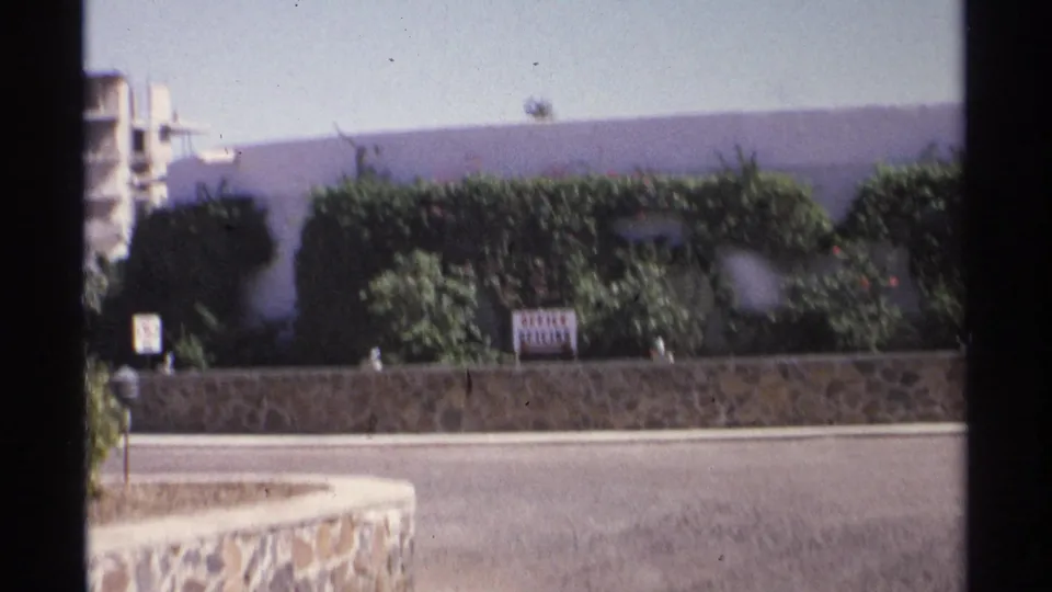 1975 Guaymas street scene with a stone wall, dense hedges, an OFFICE OFICINA sign, and a concrete building in the background.