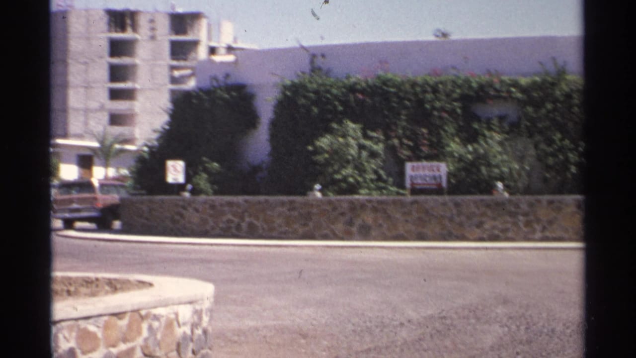 A car moves near a Guaymas roadside office frontage, with hedges, a stone wall, signage, and a concrete structure visible behind.