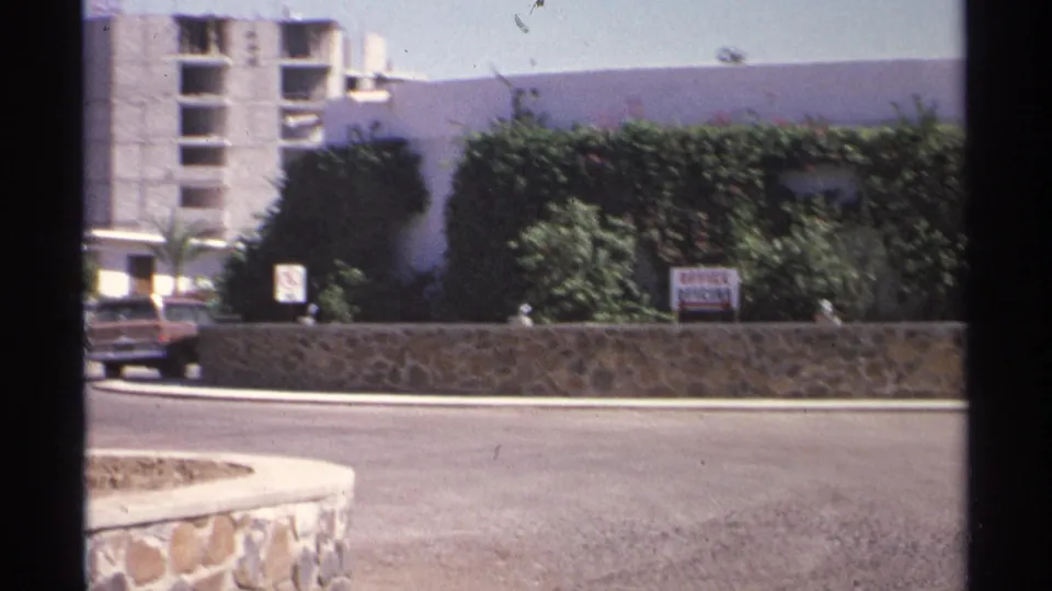 A car moves near a Guaymas roadside office frontage, with hedges, a stone wall, signage, and a concrete structure visible behind.