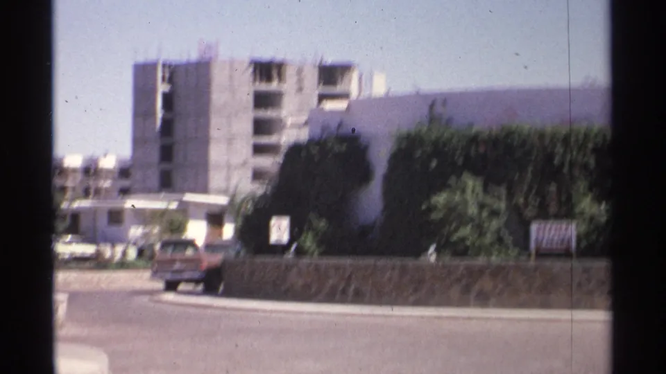Guaymas street view showing a parked or slow-moving car, stone wall, hedges, office sign, and unfinished concrete building in 1975 home-movie footage.