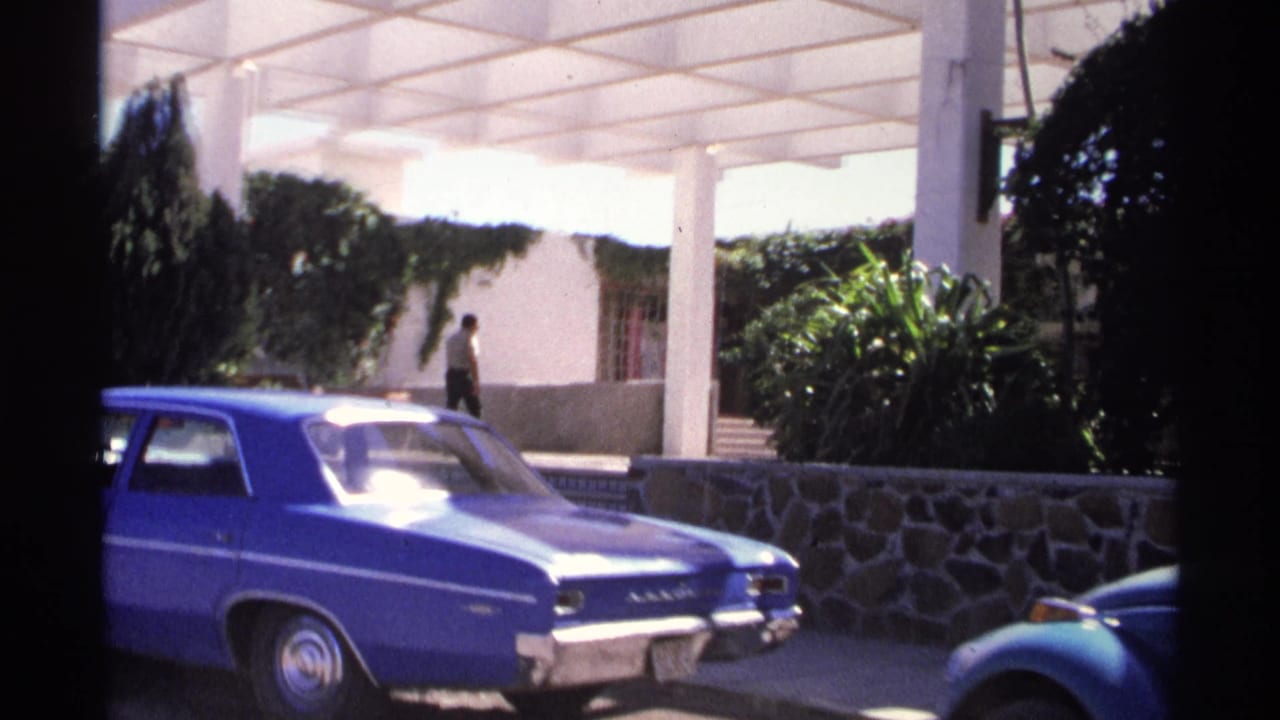 1975 Guaymas street view with a blue sedan, white pergola columns, ivy-covered wall, and a person walking near the steps.