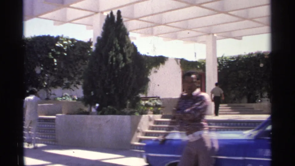 1975 Guaymas home-movie frame showing a man by a blue car, tiled steps, planted trees, and a pedestrian near an entrance.