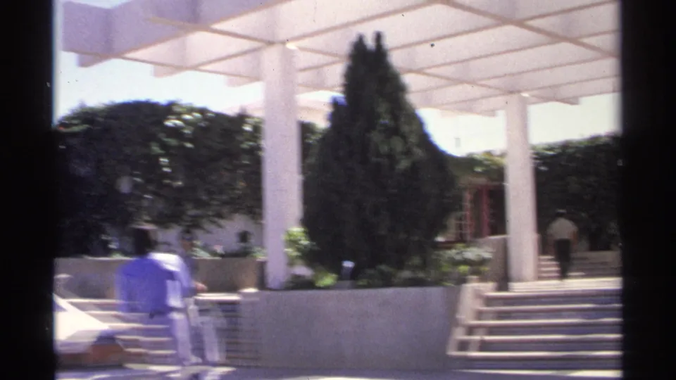 Sunlit Guaymas frame with a white pergola, tiled steps, planted trees, a pedestrian, and the rear of a blue car at the edge.
