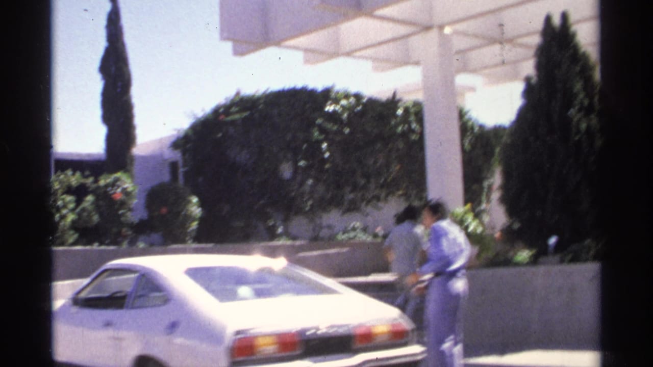 1975 Guaymas home-movie frame with two adults by a white vintage car, white columns, hedges, and tall trees in bright sun.