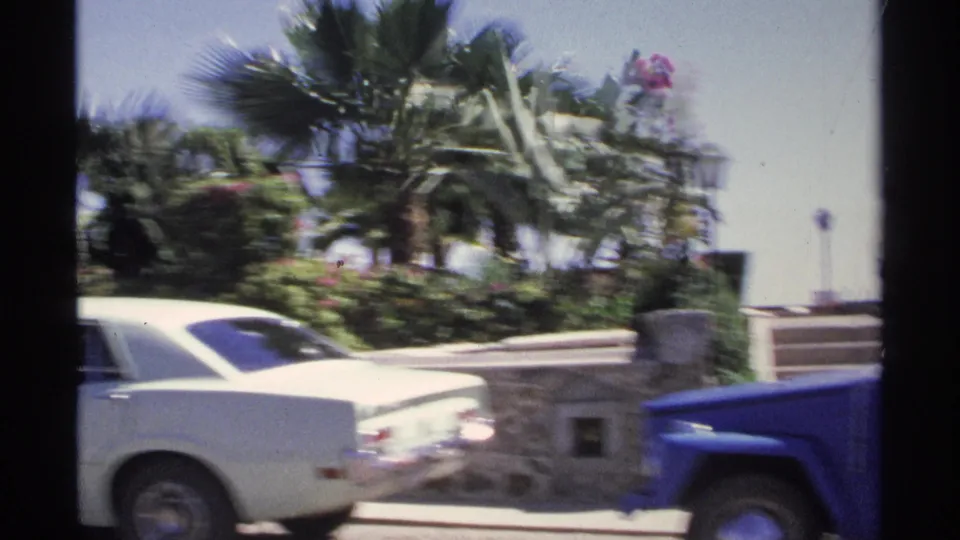 A white vintage car, blue vehicle edge, palms, flowering plants, and stonework appear in a bright 1970s Guaymas exterior view.