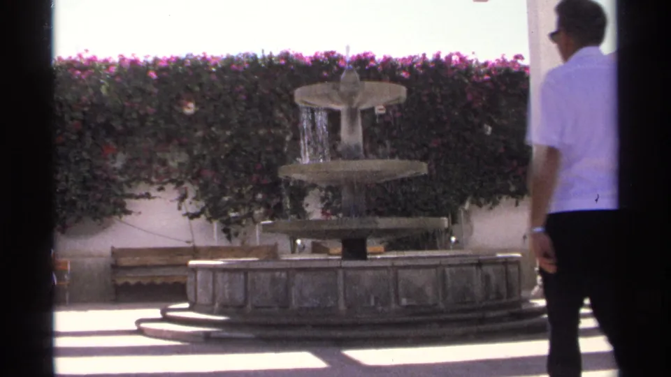 A man in a white shirt stands or walks beside a tiered stone fountain, pink flowering hedge, and paved courtyard in Guaymas, Mexico.
